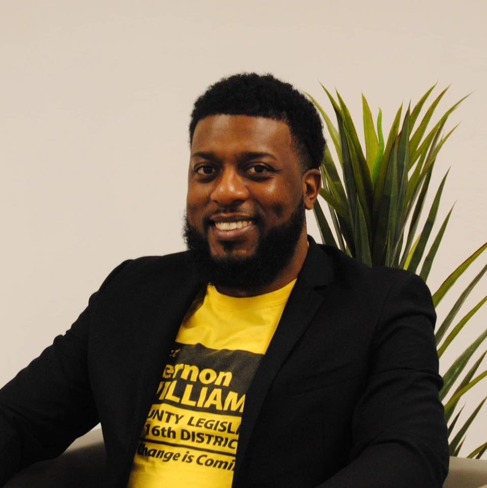Headshot of a Black man smiling in a yellow shirt and black jacket in front of a houseplant in an office.