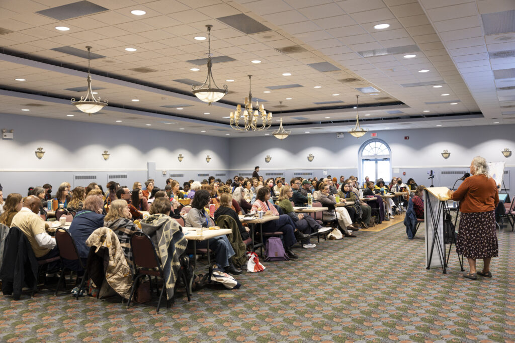 A woman in an orange shirt and patterned skirt addresses a seated crowd.